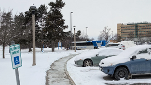 A picture of a UB Stampede Bus near a parking lot with cars that are snowed on, in which the photographer is standing on a sidewalk but not visible