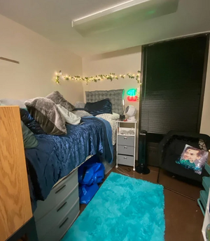 A dorm room located in the Spaulding Quadrangle of the Ellicott Complex with a ceiling light, string lights, and a raised bed with a blue blanket and pillows. The bed has storage drawers underneath it. There is a blue carpet rug in the room and the blinds on the window are closed. Next to the bed are a nightstand and a chair.
